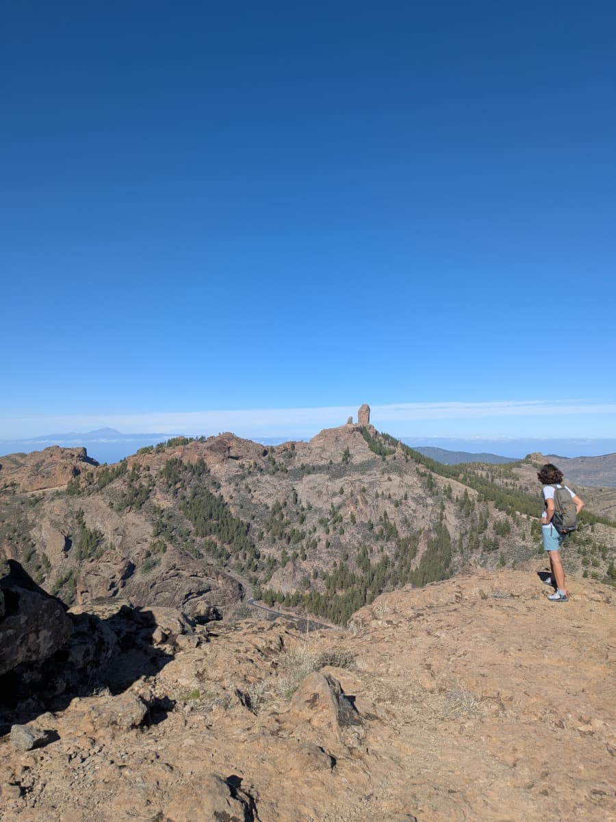 Linda steht auf einem felsigen Aussichtspunkt und blickt über eine weitläufige Berglandschaft mit dem markanten Felsen Roque Nublo im Hintergrund. Die Weite und klare Sicht auf Gran Canaria machen das Motiv typisch für Weitwandern im Winter auf den Kanaren, wo trockene Pfade, milde Temperaturen und spektakuläre Panoramen lange Etappen ermöglichen.