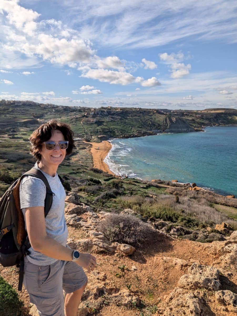 Linda steht an einem felsigen Aussichtspunkt und blickt über eine weite Ramla Bay auf Gozo mit türkisfarbenem Wasser und sanft geschwungenen Küstenlinien. Die Aufnahme unterstreicht Weitwandern im Winter auf Malta, mit klarer Sicht, ruhiger Atmosphäre und abwechslungsreichen Etappen entlang der Küste.