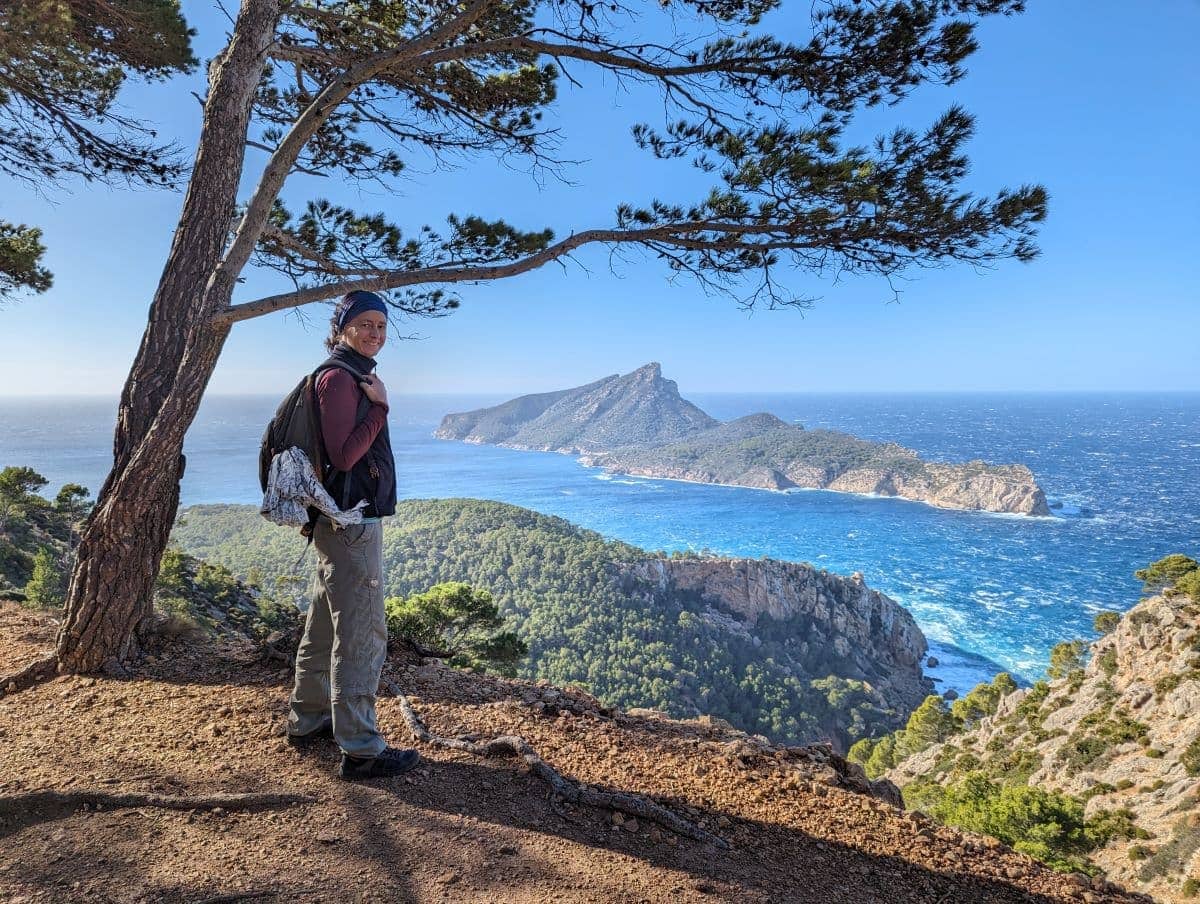 Linda steht mit Rucksack auf dem schmalen Küstenpfad GR221 unter Pinien und blickt über das tiefblaue Mittelmeer auf die zerklüftete Dracheninsel. Die Aufnahme vermittelt den Reiz von Weitwandern im Winter auf Mallorca, mit mildem Klima, klarer Fernsicht und ruhiger Stimmung abseits der Hauptsaison.
