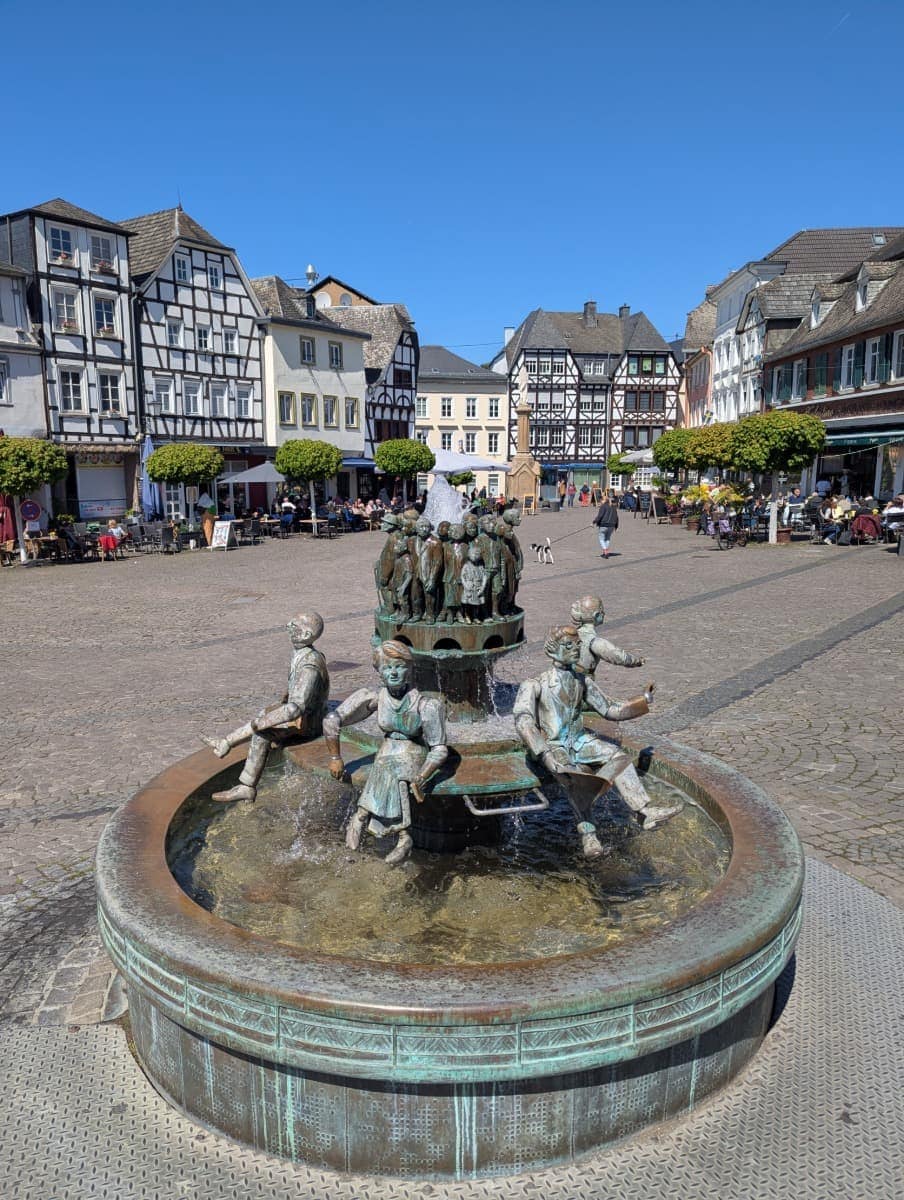 Brunnen mit bronzenen Figuren, die auf einem runden Beckenrand sitzen und scheinbar miteinander interagieren. Umgeben ist der Marktplatz von Linz am Rhein von belebten Cafés und traditionellen Fachwerkhäusern.