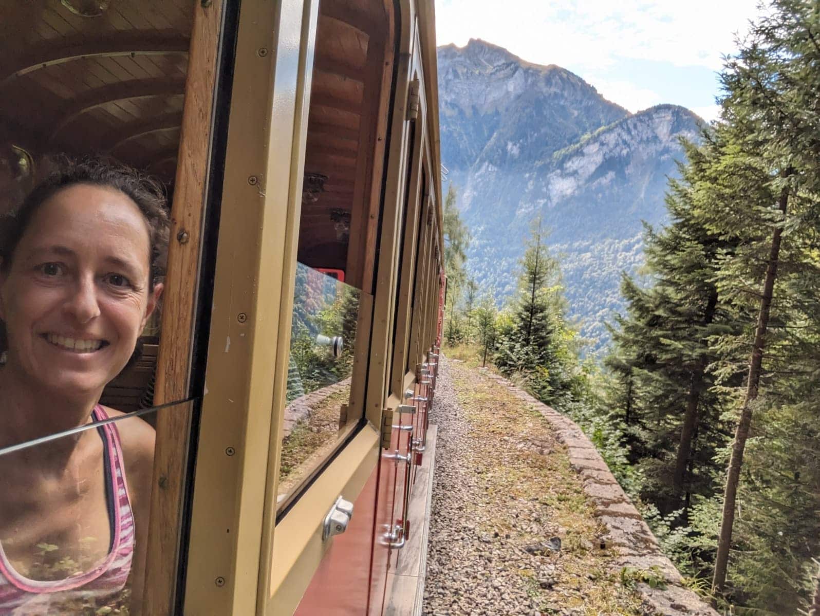 Linda lehnt lächelnd aus dem offenen Fenster eines historischen Zugwagens, während die Bahn entlang eines bewaldeten Berghangs fährt. Die Szene vermittelt nostalgisches Zugreisen in den Alpen und passt zu einem Artikel über entschleunigtes Unterwegssein mit dem Swiss Travel Pass oder Halbtax.