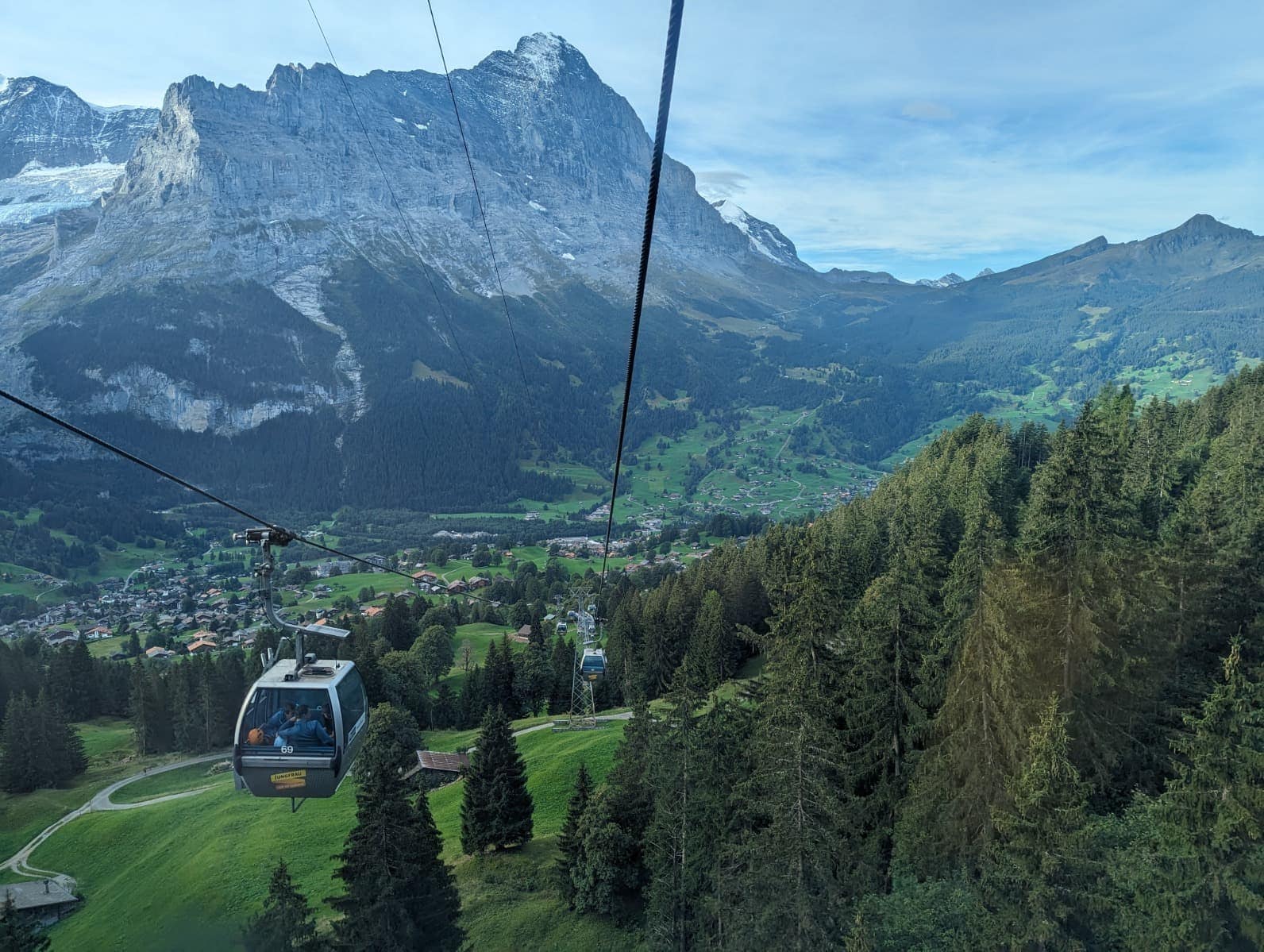 Eine Gondel schwebt über grünen Wiesen und dichten Wäldern mit Blick auf ein Dorf und ein markantes Bergmassiv im Hintergrund. Das Panorama zeigt, wie gut sich Bahn- und Bergbahnfahrten in der Schweiz mit dem Swiss Travel Pass oder Halbtax kombinieren lassen.