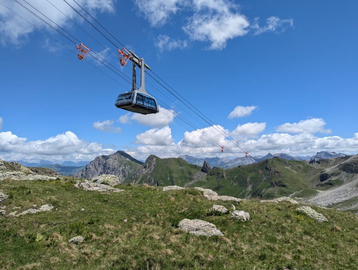 Eine moderne Seilbahnkabine fährt hoch über alpine Grasflächen und schroffe Gipfel unter blauem Himmel. Das Bild unterstreicht die spektakulären Aussichten, die Reisende bei Bergfahrten mit Swiss Travel Pass oder Halbtax erwarten.