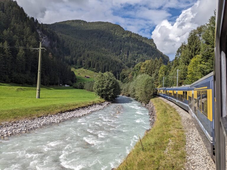 Ein Zug fährt entlang eines türkisfarbenen Flusses durch ein enges Tal mit Wiesen und bewaldeten Hängen. Diese typische Schweizer Landschaft macht deutlich, warum Bahnreisen mit Swiss Travel Pass oder Halbtax ein Highlight jeder Reise sind.
