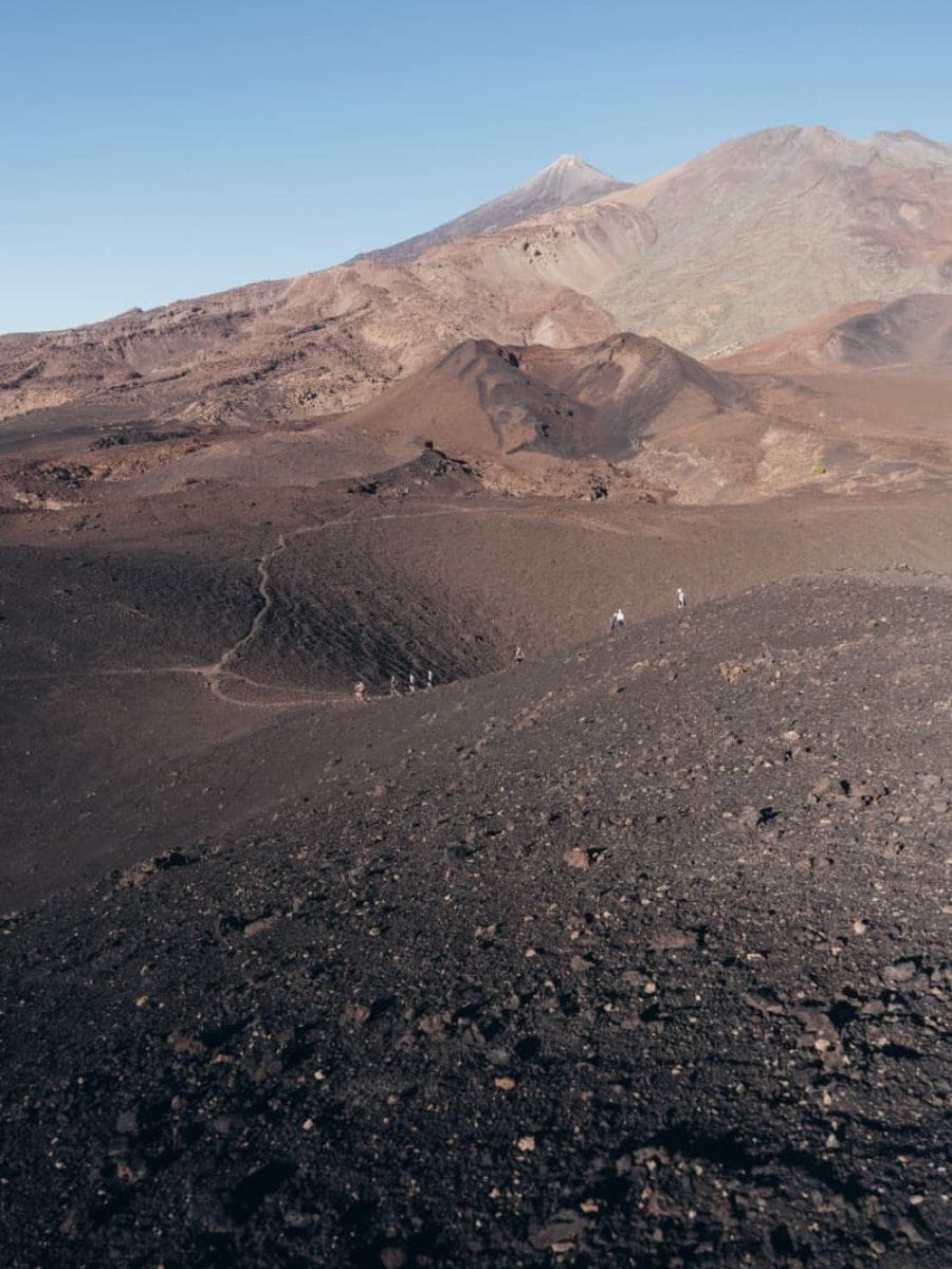 Eine kleine Gruppe von Wanderern zieht über einen schmalen Pfad durch eine karge Vulkanlandschaft aus schwarzem Lavagestein mit weiten Bergen im Hintergrund. Das Motiv unterstreicht Weitwandern im Winter auf den Kanaren, wo milde Temperaturen und einsame Routen durch ursprüngliche Landschaften lange Etappen ermöglichen.