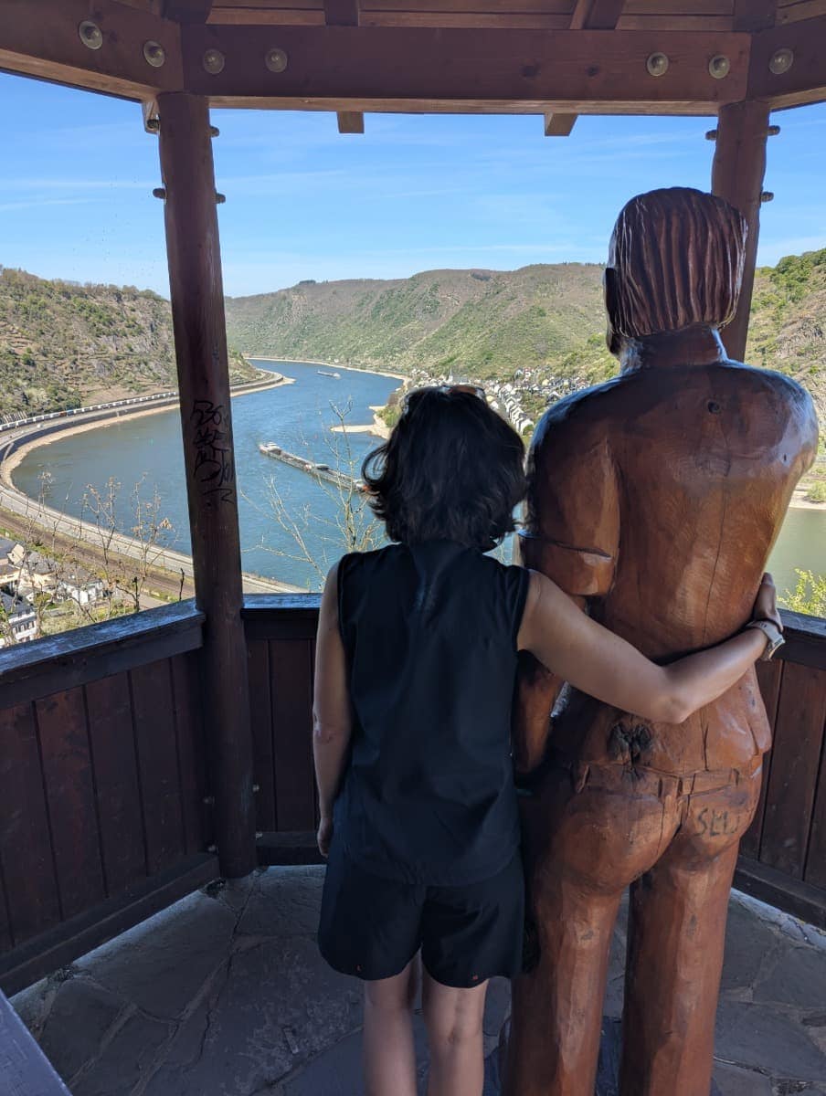 Linda steht auf der Europakanzel am RheinBurgenWeg neben einer Holzfigur und blickt über die große Rheinschleife des Rheins bei Boppard ins Mittelrheintal. Die überdachte Aussichtsplattform bietet eines der bekanntesten Panoramen entlang dieser Rheinsteig Alternative.