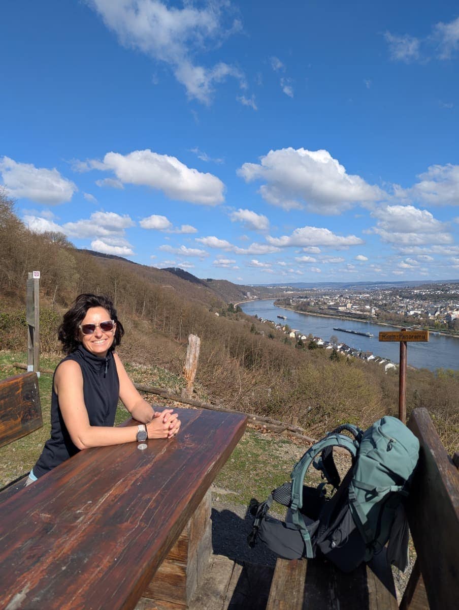 Linda sitzt an einem Holztisch auf einem Aussichtspunkt am RheinBurgenWeg, einer Alternative zum Rheinsteig, mit weitem Blick über das beeindruckende Rheinpanorama oberhalb von Koblenz.