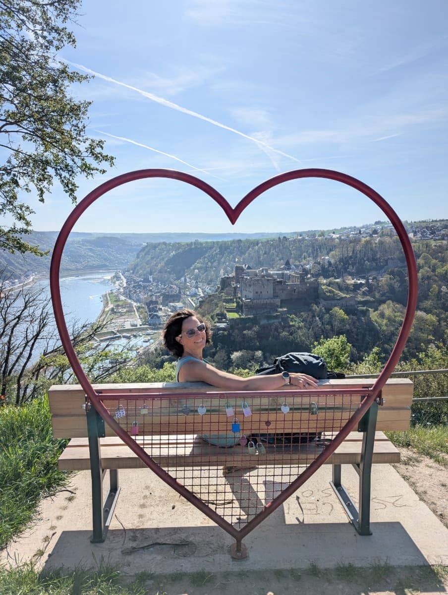 Linda sitzt auf einer Bank im Rheinherz am RheinBurgenWeg mit Blick über den Rhein auf St. Goar und die Burg Rheinfels im Mittelrheintal.