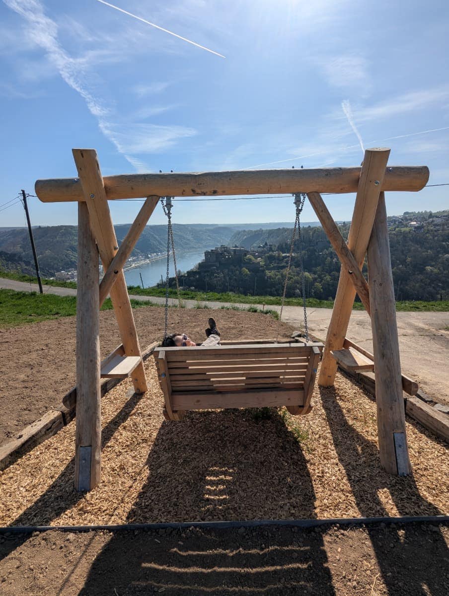 Linda liegt auf einer großen Holzschaukel an einem Aussichtspunkt am RheinBurgenWeg mit Blick über den Rhein und die Burg Rheinfels oberhalb von St. Goar.