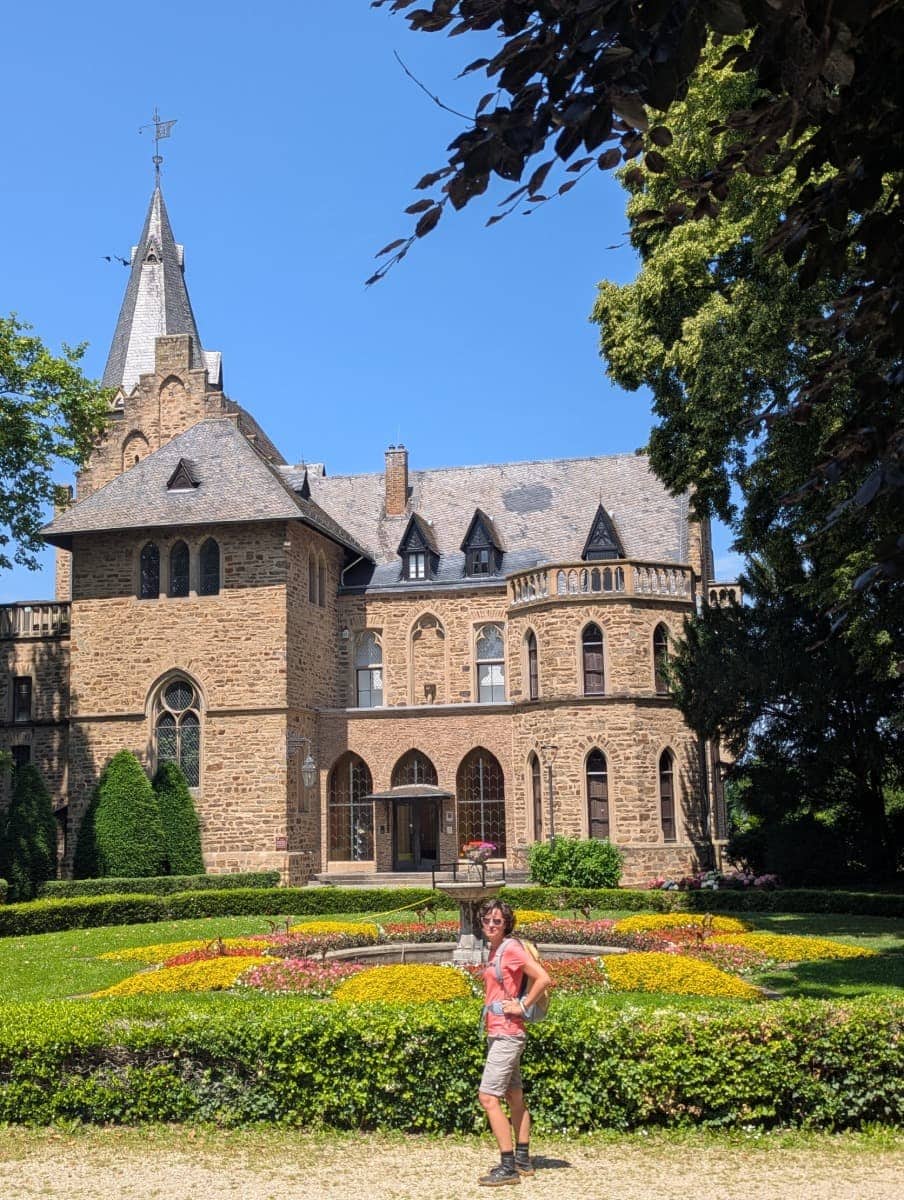 Linda steht im Garten von Schloss Sinzig am RheinBurgenWeg vor dem neugotischen Schlossbau mit Türmen, Sandsteinfassade und gepflegten Blumenbeeten.