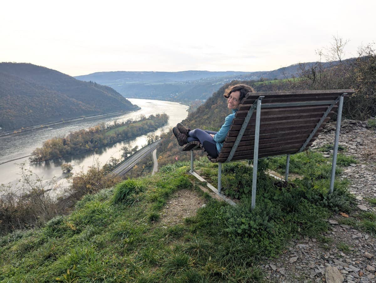 Linda sitzt auf einer Bank am RheinBurgenWeg oberhalb von Bacharach und blickt über das weite Rheintal und den geschwungenen Fluss im UNESCO-Welterbe Oberes Mittelrheintal.