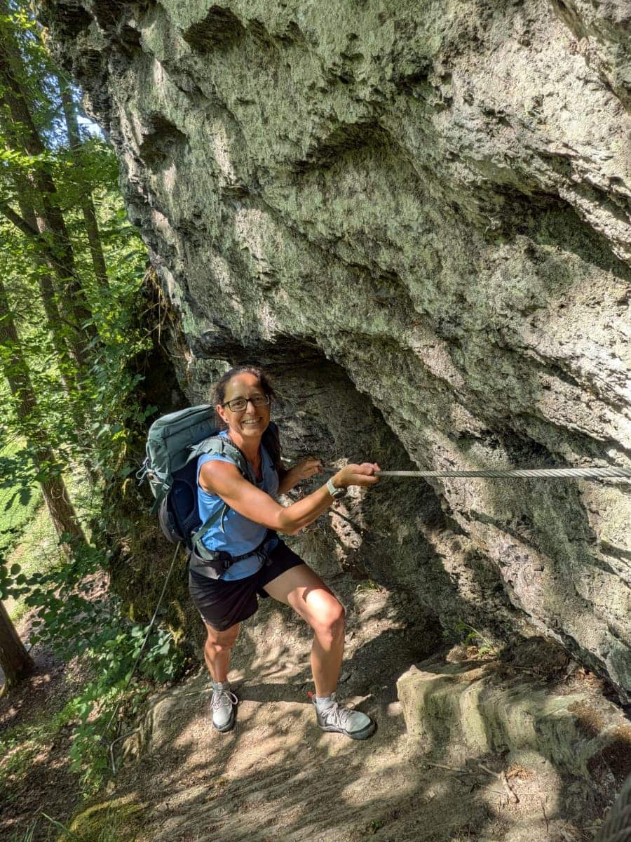 Linda hält sich an einem Drahtseil entlang eines schmalen Felswegs am Diellaysteig fest und meistert eine der anspruchsvolleren Passagen durch die bewaldeten Hänge auf dem Saar Hunsrück Steig.