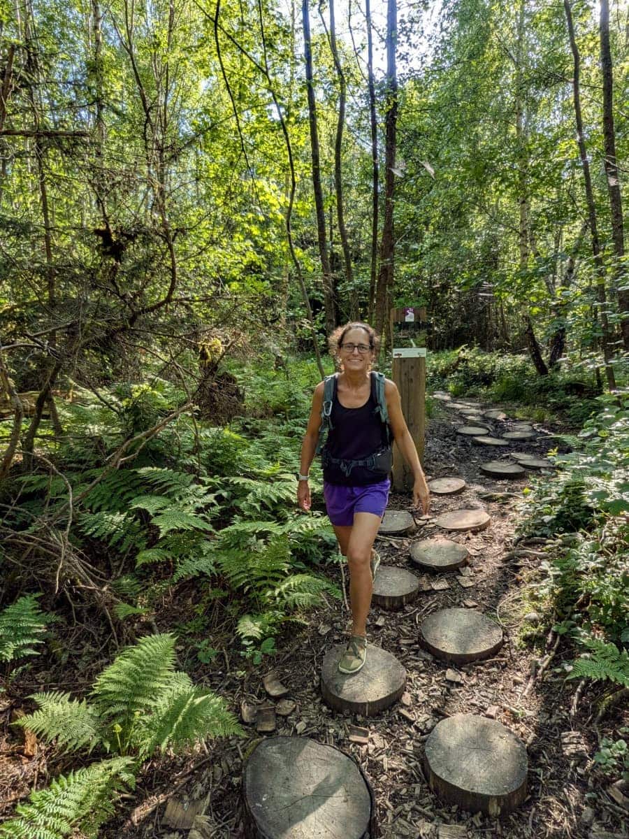 Linda wandert in Kastellaun auf einem naturbelassenen Waldpfad der Traumschleife Burgstadt-Pfad über runde Holztrittsteine durch einen von Farnen und dichtem Grün geprägten Abschnitt des Hunsrück.