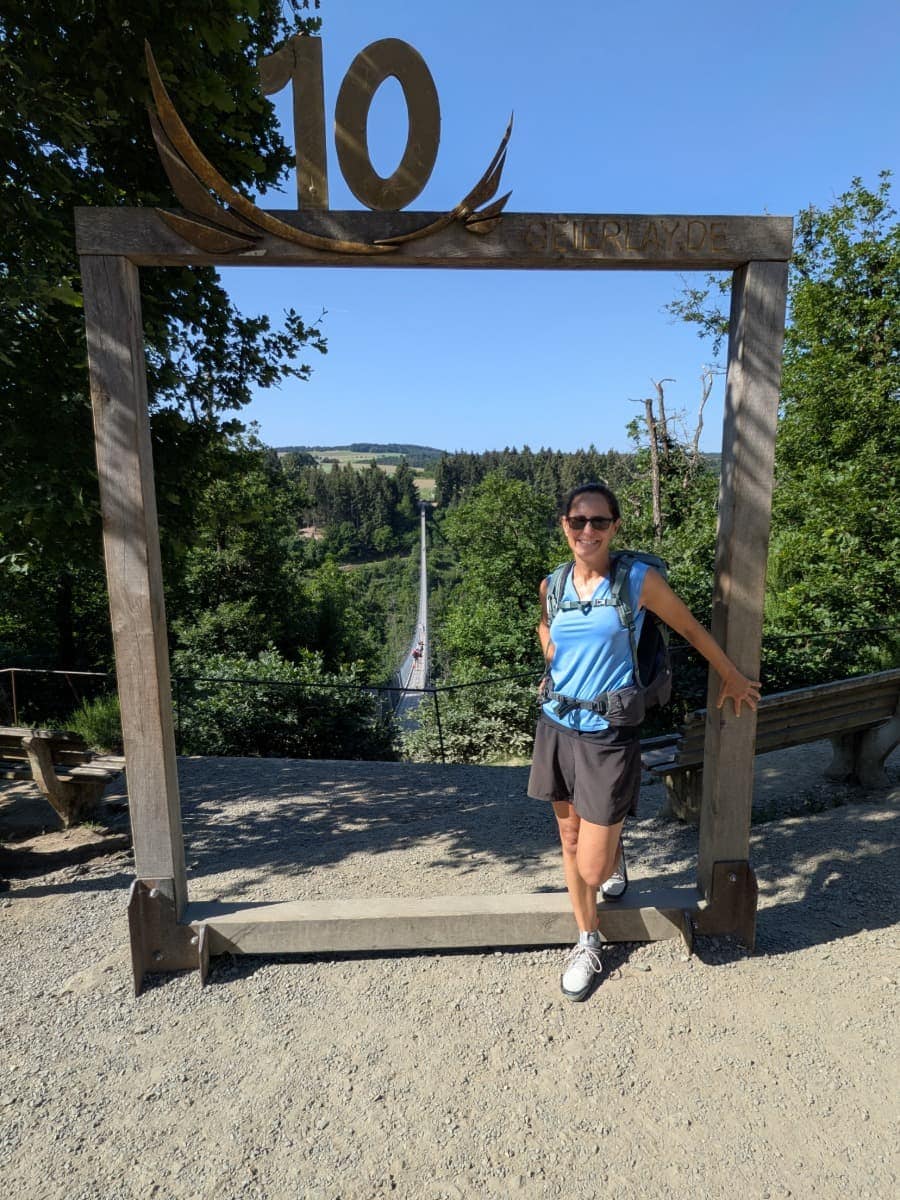 Linda steht am Aussichtspunkt der Geierlay und posiert vor dem Bilderrahmen mit Blick auf die Hängeseilbrücke, die sich durch das bewaldete Tal im Hunsrück spannt.