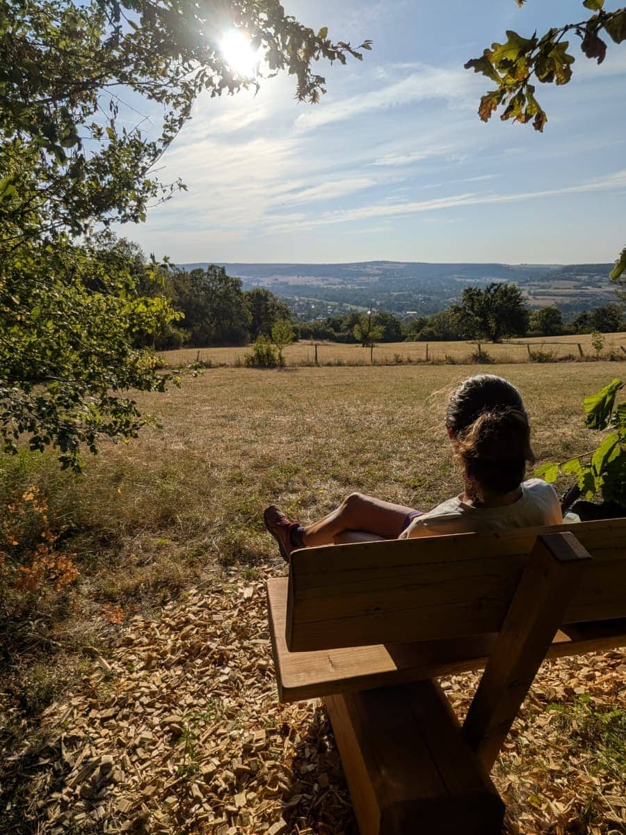Linda sitzt auf einer Holzbank und blickt über die sanfte Hügellandschaft des Bliesgaus entlang des Bliessteigs.
