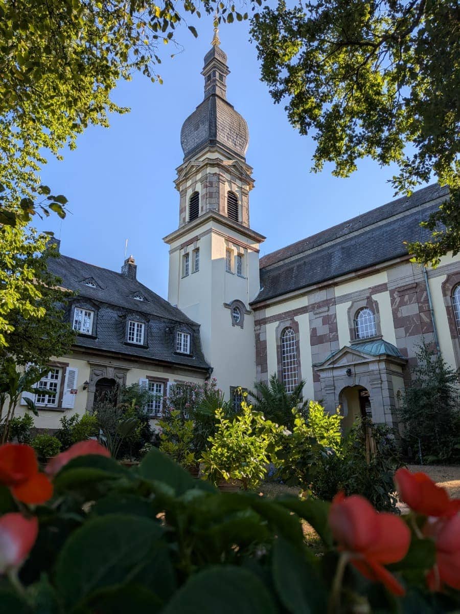 Historische Kloster- und Kirchengebäude mit markantem Zwiebelturm stehen zwischen Bäumen und Blumen in Blieskastel am Bliessteig.