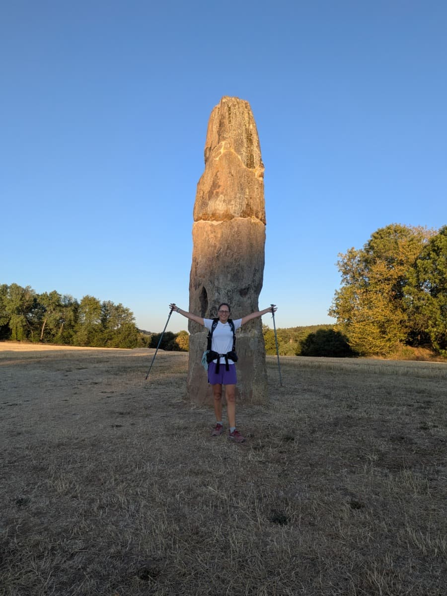 Linda steht mit ausgebreiteten Trekkingstöcken vor dem rund 6,5 Meter hohen Gollenstein bei Blieskastel, einem markanten Menhir an der vierten Etappe des Bliessteigs im Saarland.