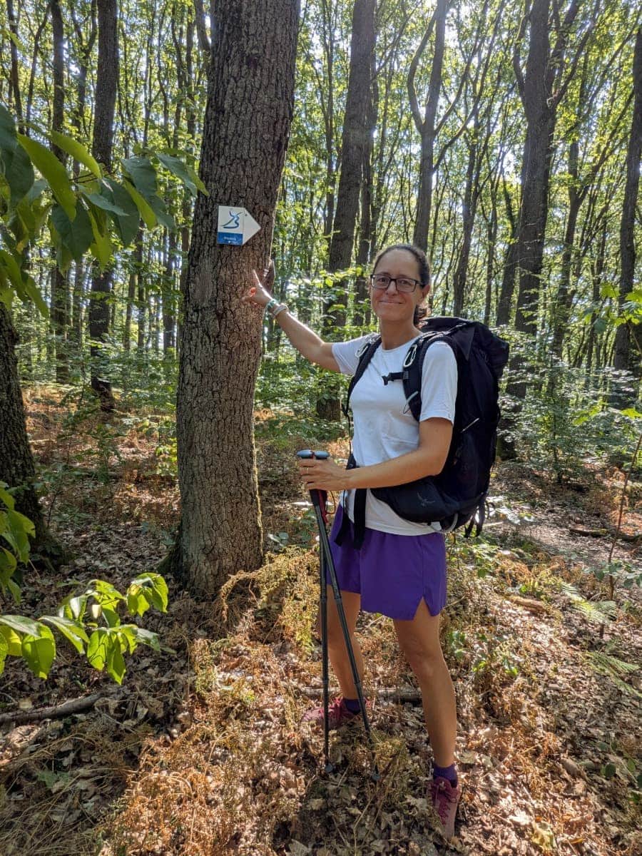 Linda steht mit Rucksack und Trekkingstöcken neben der Wegmarkierung des Bliessteigs in einem schattigen Waldstück.