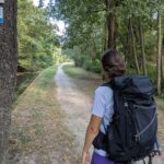 Linda wandert mit großem Rucksack auf einem schattigen Weg entlang eines Kanals, während die Bliessteig Wegmarkierung am Baum den Verlauf der Fernwanderung weist.
