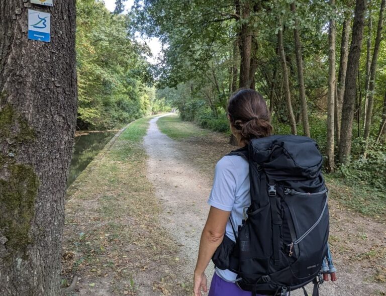 Linda wandert mit großem Rucksack auf einem schattigen Weg entlang eines Kanals, während die Bliessteig Wegmarkierung am Baum den Verlauf der Fernwanderung weist.