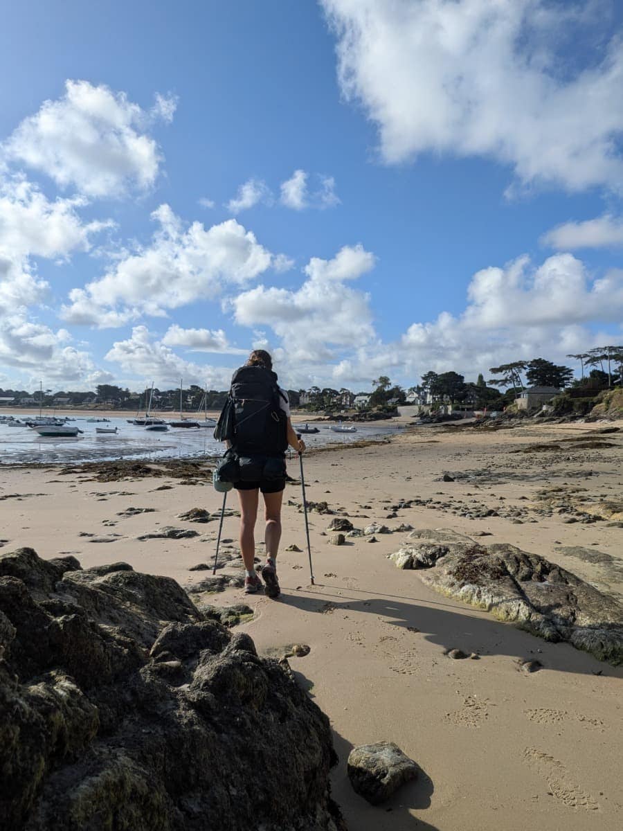 Linda wandert mit Trekkingstöcken über den Sandstrand der Bucht von Rothéneuf bei Ebbe auf dem Zöllnerpfad GR34 entlang der bretonischen Küste.