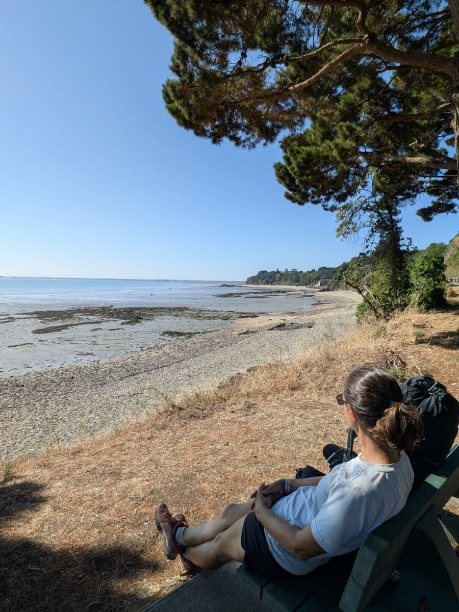 Linda genießt eine Pause mit Blick auf den ruhigen Strand und das Meer während ihrer Wanderung auf dem Zöllnerweg.