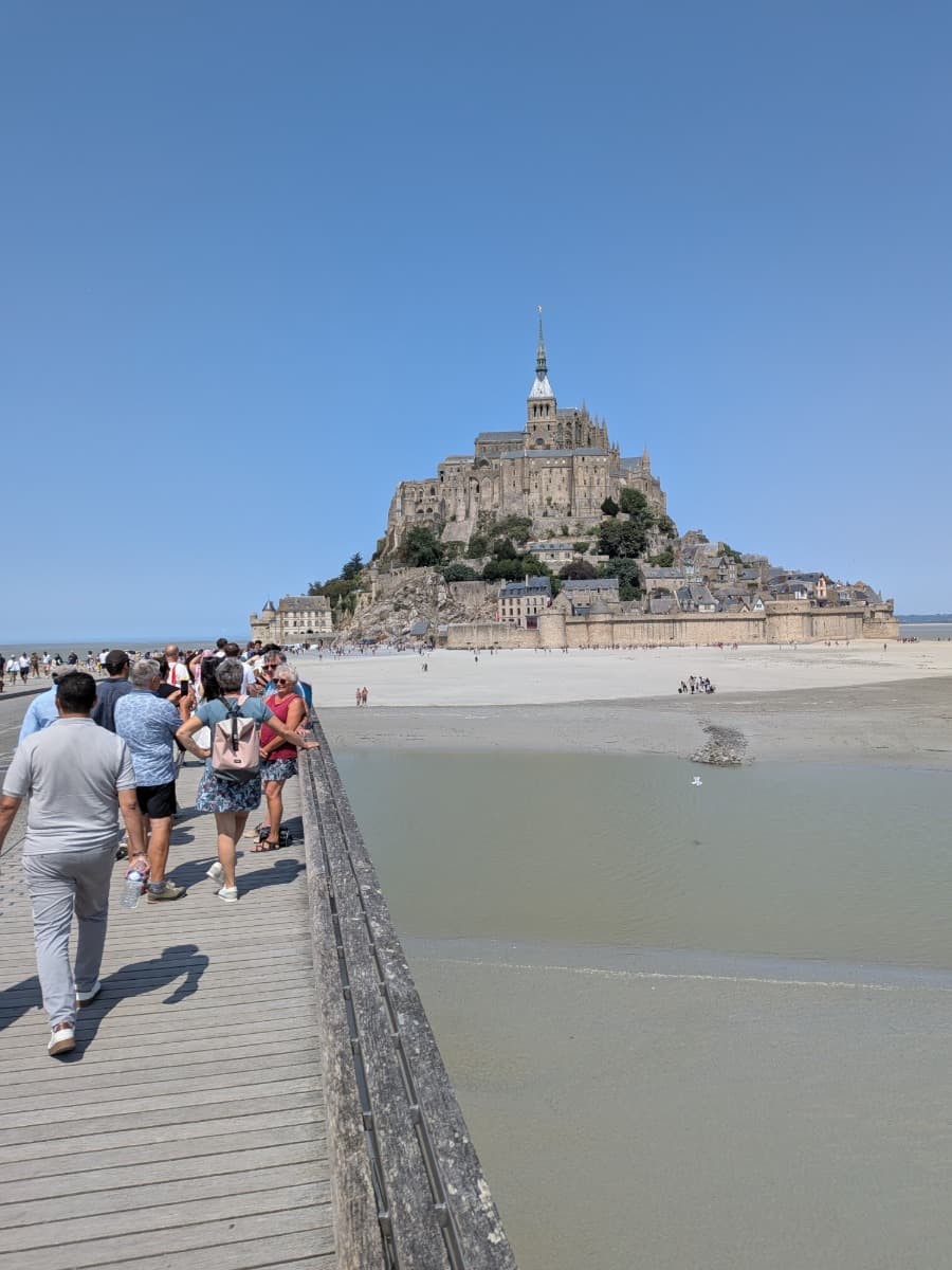 Besucher überqueren den Passerelle du Mont-Saint-Michel, während die Klosterinsel mit der Abtei eindrucksvoll vor blauem Himmel aufragt.