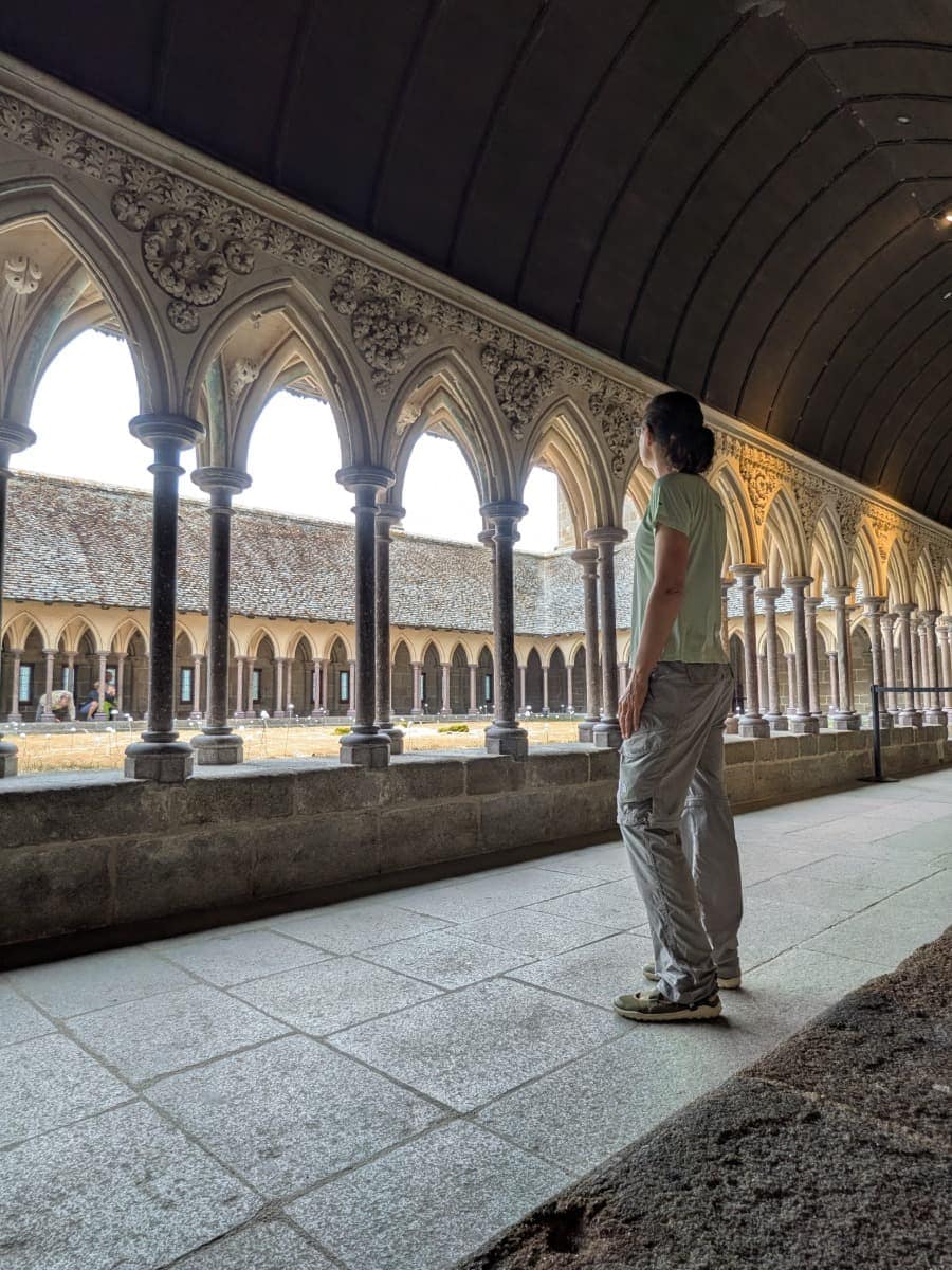 Linda betrachtet den historischen Kreuzgang der Abtei Mont-Saint-Michel mit seinen doppelten Säulenreihen und dem begrünten Innenhof.