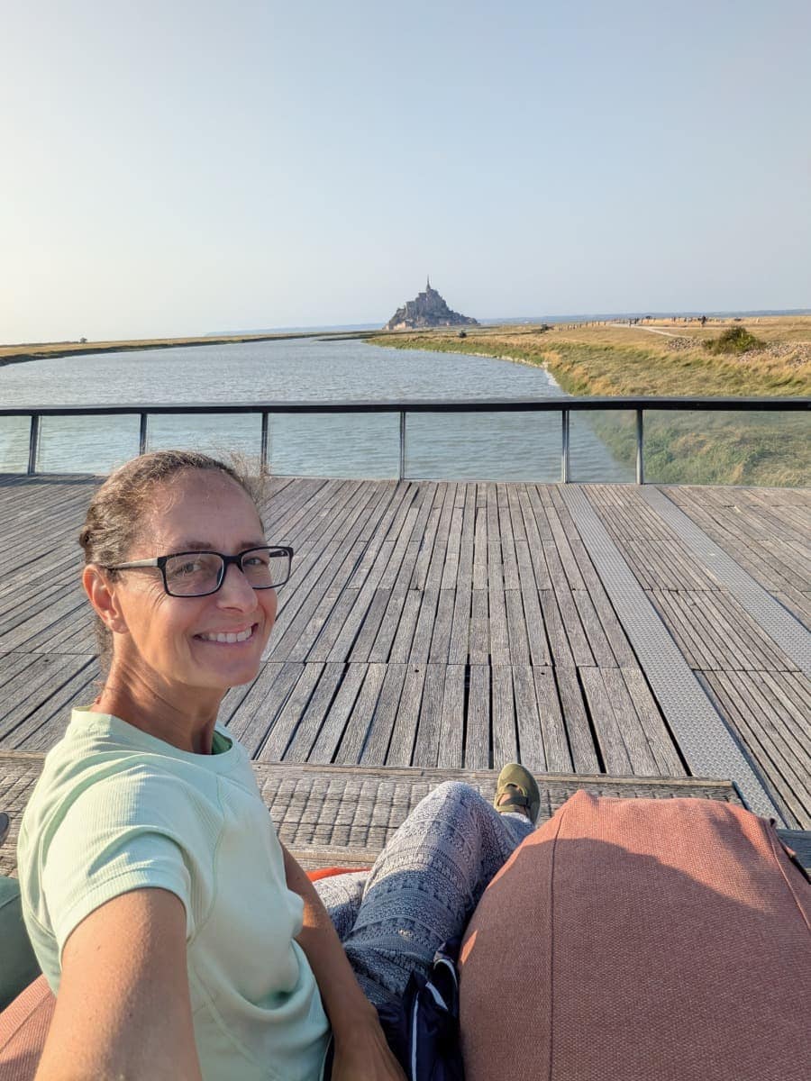 Linda entspannt auf der Terrasse des Barrage du Mont-Saint-Michel mit Blick über den Fluss Cuesnon und den Mont-Saint-Michel, der in der Ferne aus der weiten Bucht aufragt.