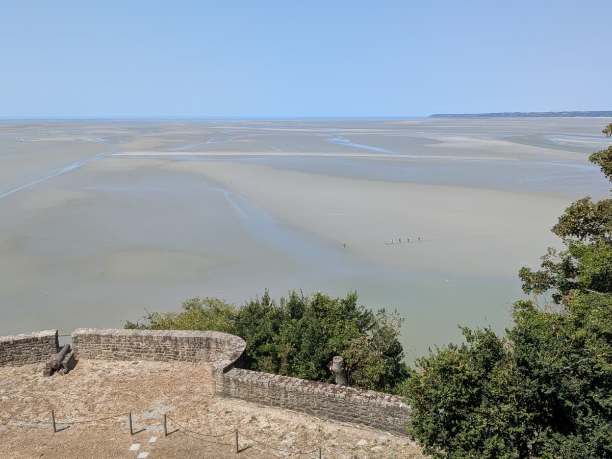 Blick von den Wehrmauern des Mont-Saint-Michel über das weitläufige Watt der Baie du Mont-Saint-Michel bei Ebbe, in der eine Gruppe eine Wattwanderung unternimmt.