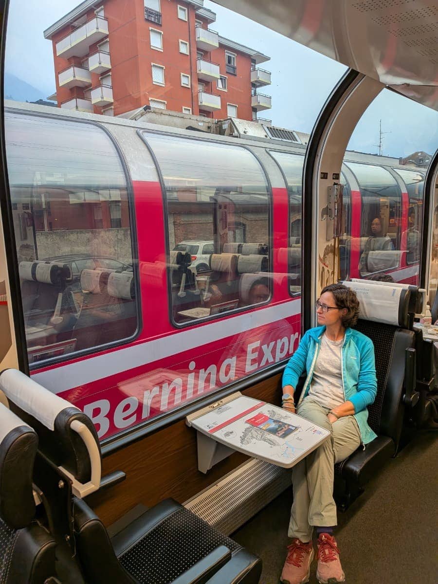 Linda sitzt im Panoramawagen des Bernina Express und blickt am Bahnhof Tirano auf einen gegenüberstehenden roten Zug der Rhätischen Bahn, dessen große Fenster und Schriftzug sich direkt neben ihrem Platz spiegeln.