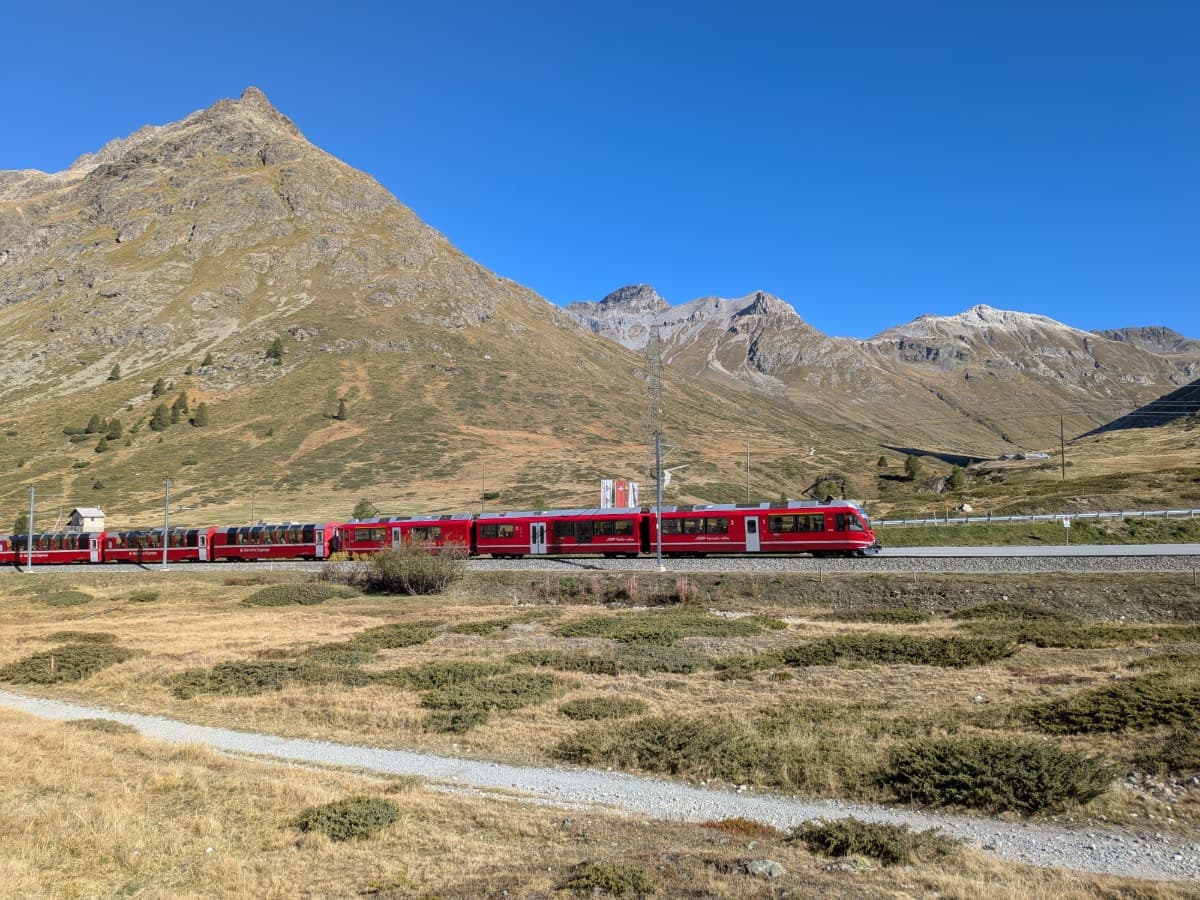 Der Bernina Express fährt über den Berninapass durch die offene Hochgebirgslandschaft, eingerahmt von kargen Hängen und markanten Gipfeln unter tiefblauem Himmel.