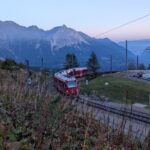 Zwei rote Züge der Rhätischen Bahn begegnen sich auf den Gleisen bei Alp Grüm, hoch über dem Val Poschiavo und vor den markanten Bergen der Berninagruppe im Abendlicht.
