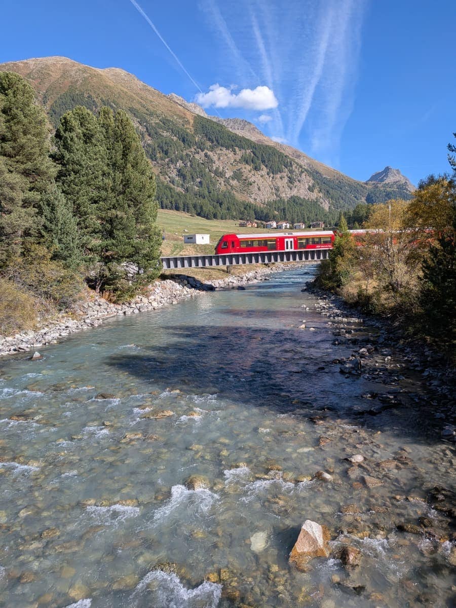Der Bernina Express überquert eine Brücke über die Ova da Bernina, während sich dahinter sonnige Hänge und die Berglandschaft rund um den Berninapass öffnen.