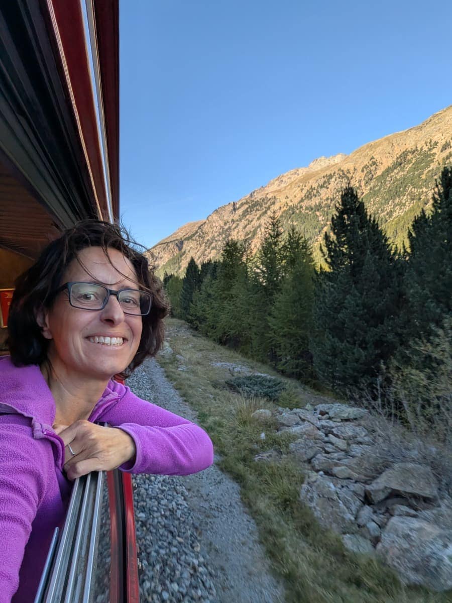 Linda lehnt lächelnd aus dem offenen Fenster eines roten Regionalzuges der Rhätischen Bahn und genießt die Fahrt durch die bewaldete Berglandschaft entlang der Berninalinie.