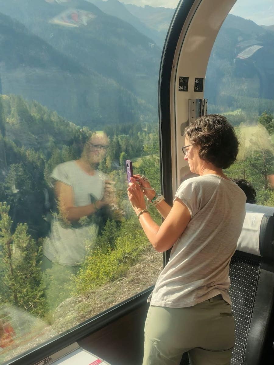 Linda fotografiert im Bernina Express durch das hohe Panoramafenster die sonnige Berglandschaft, während sich ihr Spiegelbild im Glas mit den bewaldeten Hängen draußen überlagert.