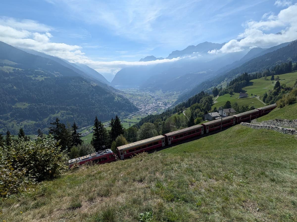Der Bernina Express fährt oberhalb von Poschiavo durch die grünen Hänge des Val Poschiavo, während sich tief im Tal der Ort und dahinter die markanten Bergketten des Puschlavs ausbreiten.