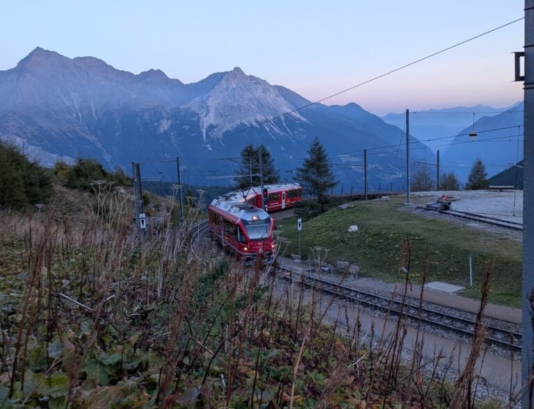 Zwei rote Züge der Rhätischen Bahn begegnen sich auf den Gleisen bei Alp Grüm, hoch über dem Val Poschiavo und vor den markanten Bergen der Berninagruppe im Abendlicht.