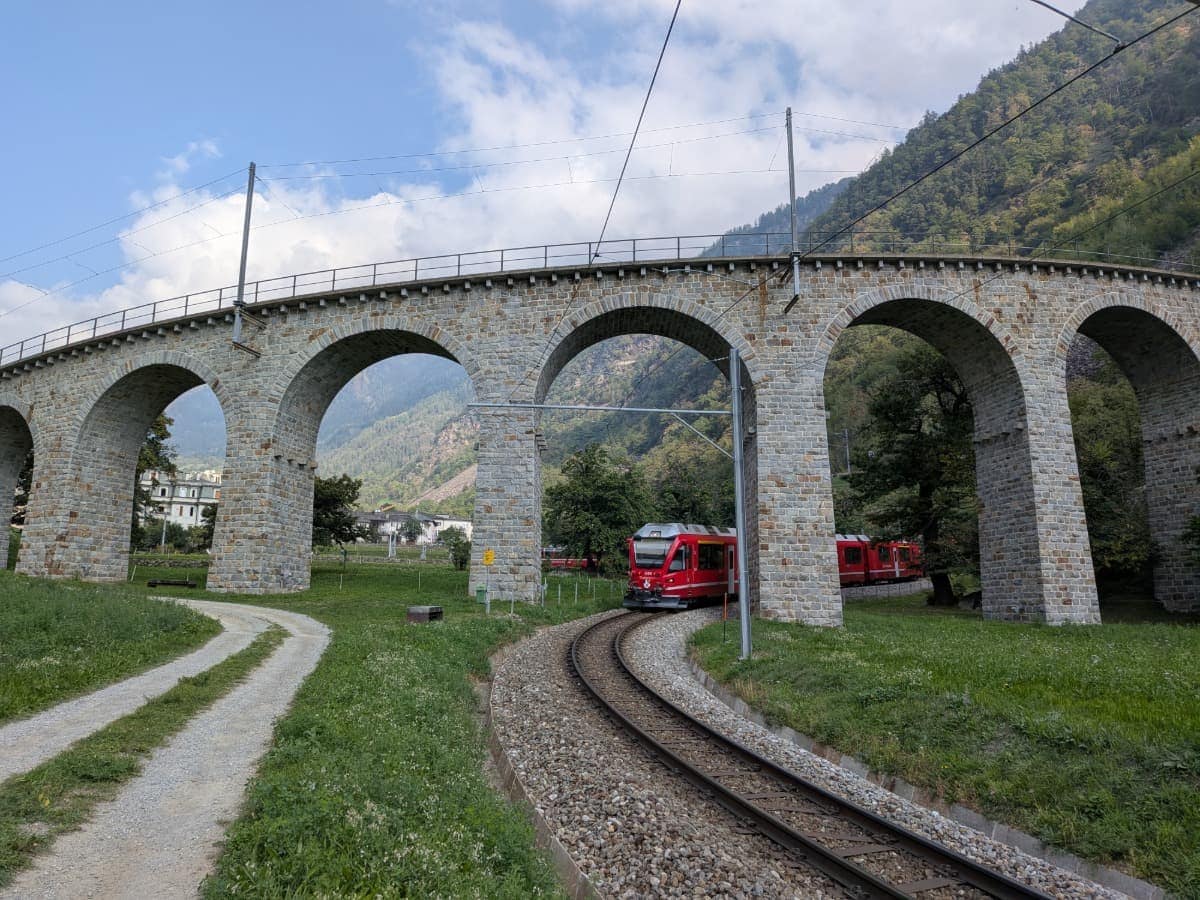Der roter Zug Bernina Express fährt durch die enge Kurve unter dem historischen Kreisviadukt von Brusio, dessen gemauerte Bögen sich vor den bewaldeten Hängen des Val Poschiavo erheben.