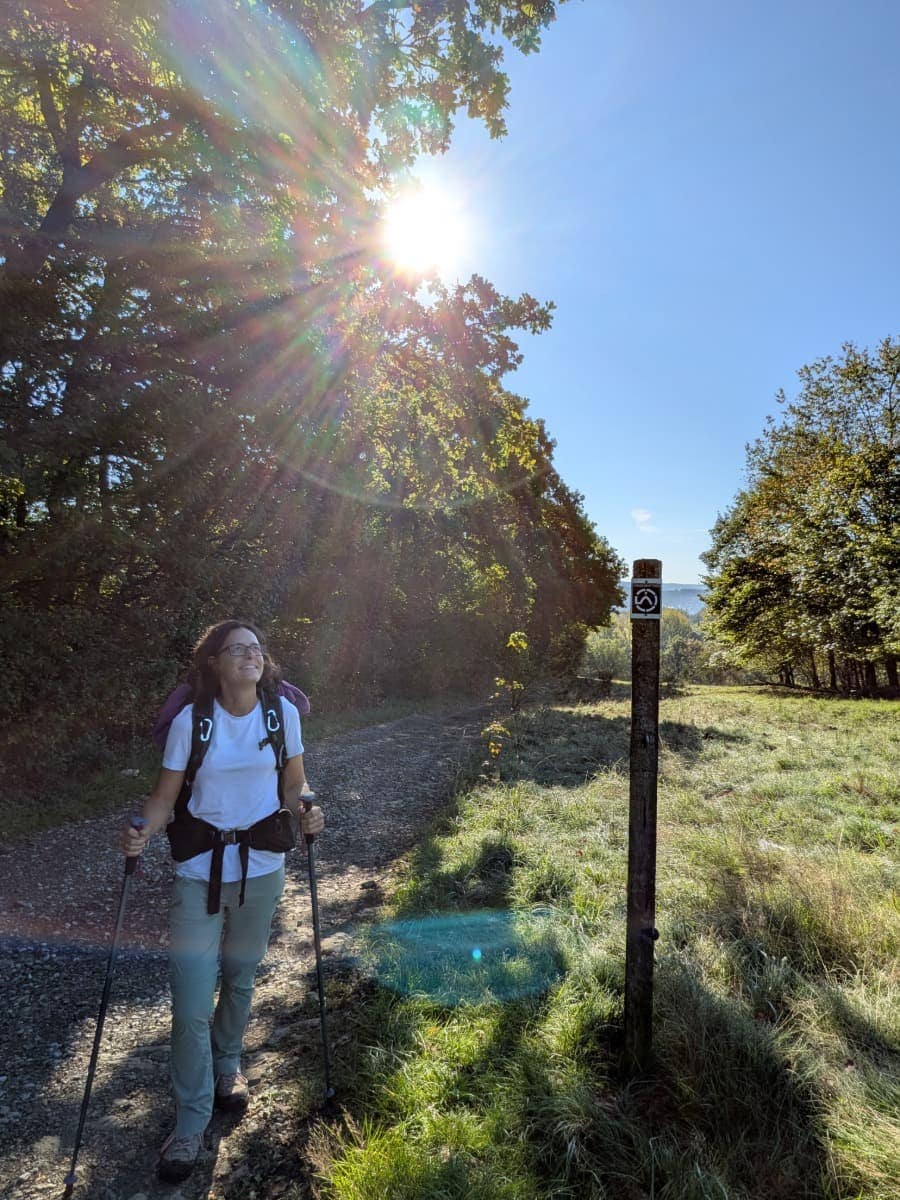 Linda steht mit Rucksack und Wanderstöcken neben einer Wegmarkierung des Druidensteigs, während Sonnenstrahlen durch die Baumkronen fallen.