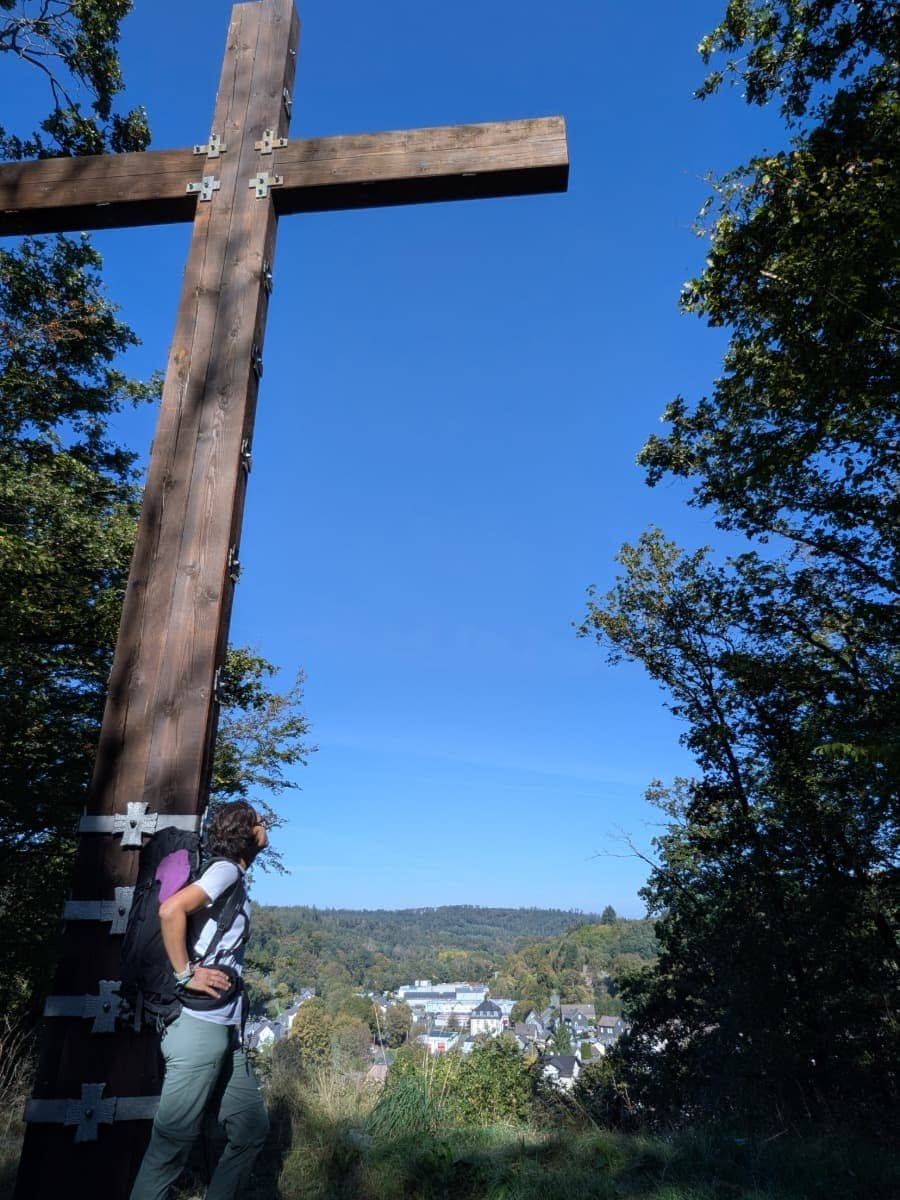 Linda steht mit Wanderrucksack am großen hölzernen Friedenskreuz Wehbach mit Blick auf die Häuser und bewaldeten Hügel im Siegtal.