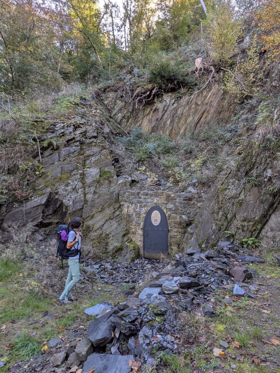 Linda steht mit Wanderrucksack vor dem verschlossenen Stolleneingang des ehemaligen Kobalterzbergwerks Alexander im Westerwald, umgeben von Felsen und Wald.