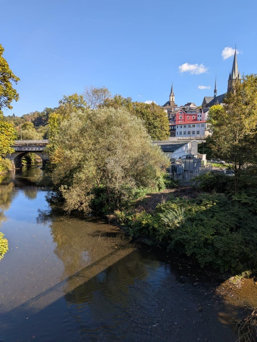 Die Sieg fließt durch Kirchen unter einer Brücke hindurch, während dahinter Häuser und Kirchtürme zwischen herbstlichen Bäumen aufragen.