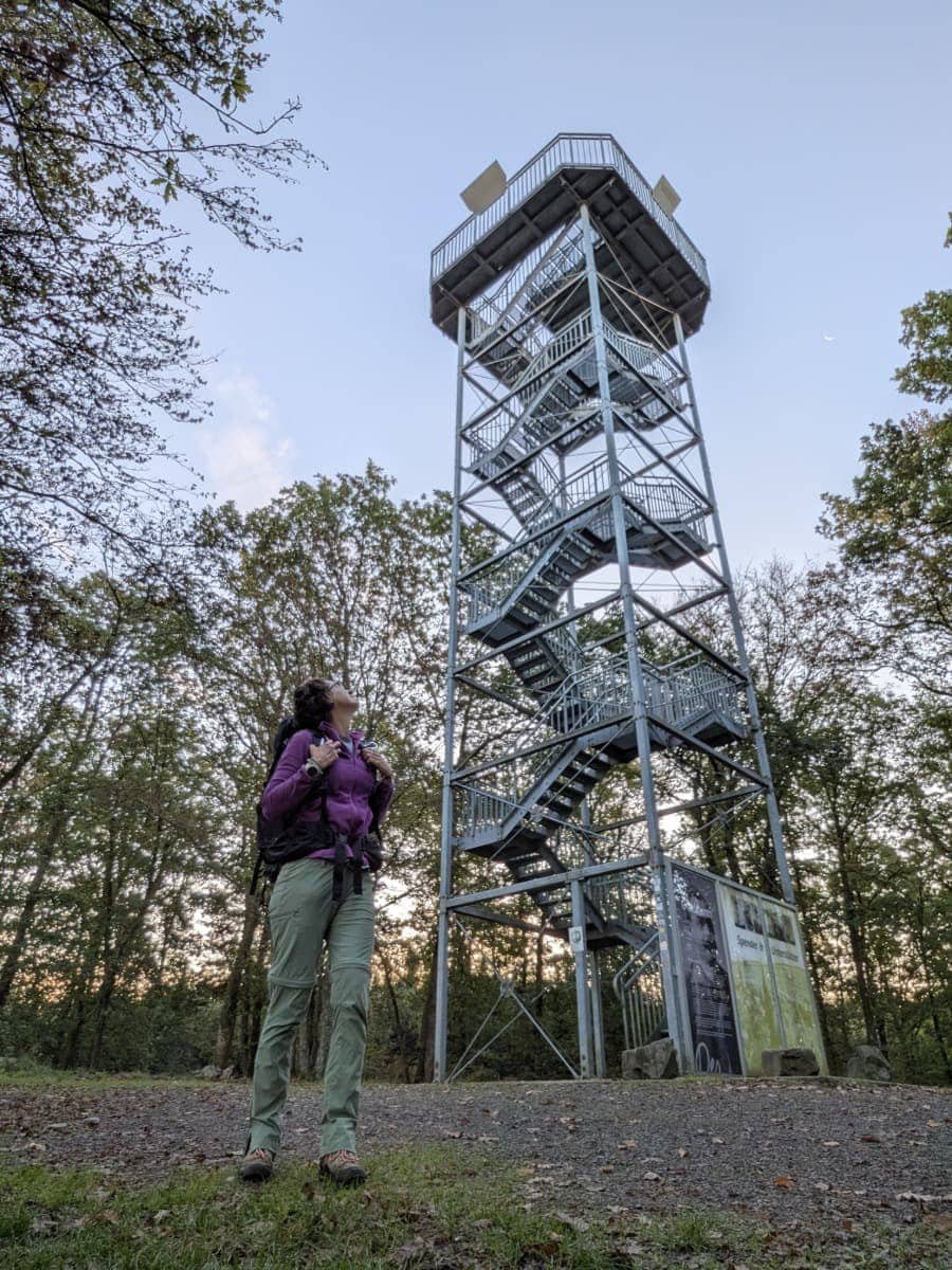 Linda steht mit Wanderrucksack vor dem stählernen Ottoturm am Druidensteig im Westerwald und blickt zur Aussichtsplattform hinauf.