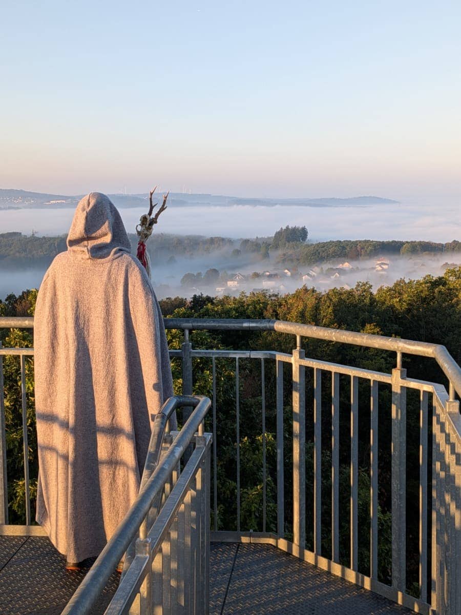 Ein Druide im grauen Kapuzenumhang blickt vom Ottoturm über bewaldete Hügel und ein von Morgennebel bedecktes Tal im Westerwald.