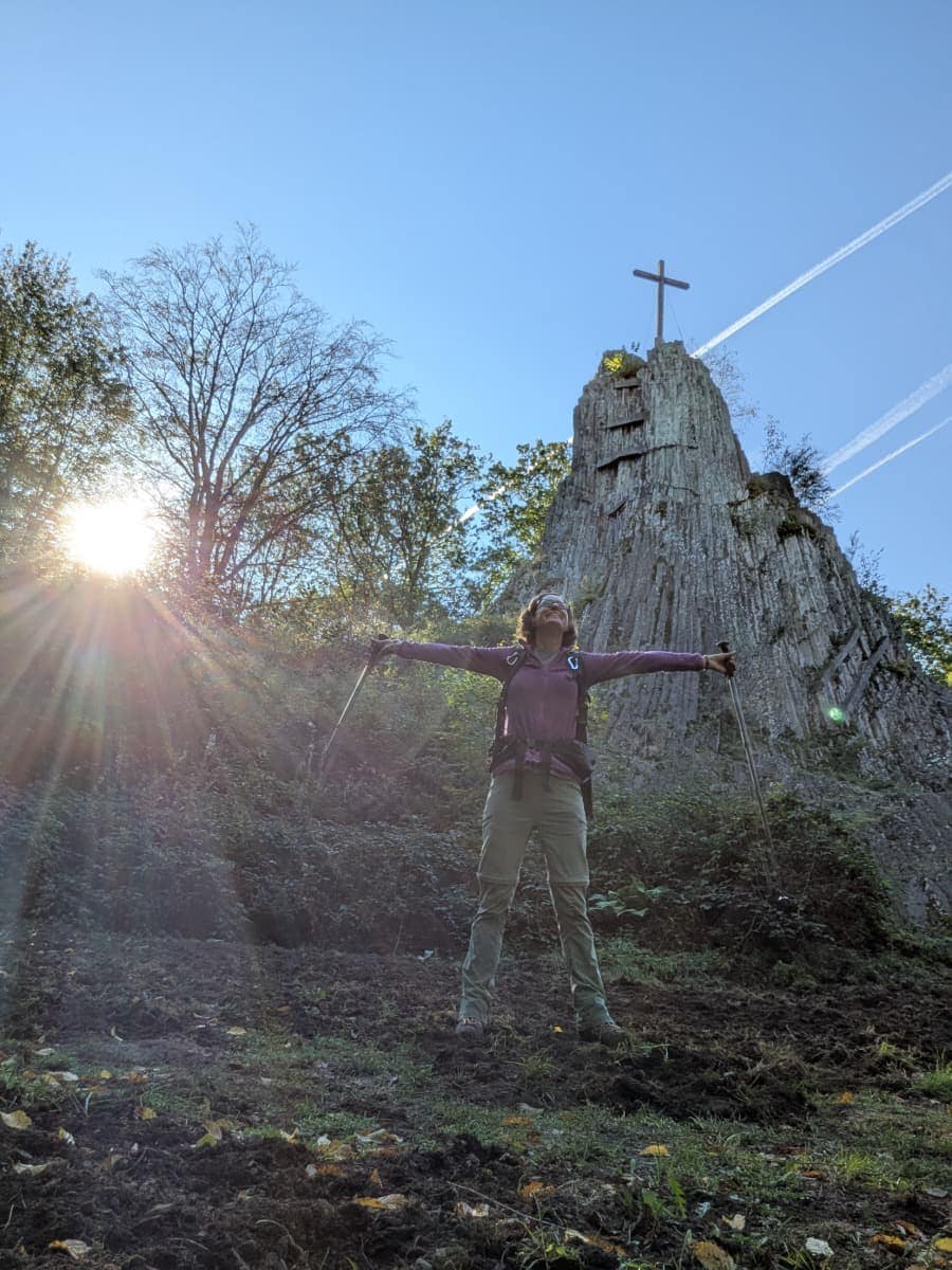 Auf ihrer Mehrtagestour im Westerwald steht Linda mit ausgebreiteten Armen und Wanderstöcken vor dem Druidenstein mit seinem Gipfelkreuz.