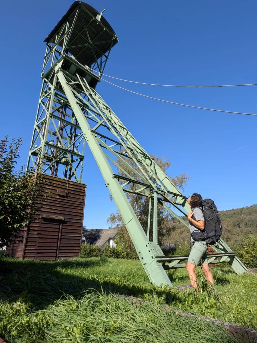 Linda blickt mit ihrem großen Wanderrucksack zum grünen Förderturm am Bergbaumuseum in Herdorf-Sassenroth hinauf.