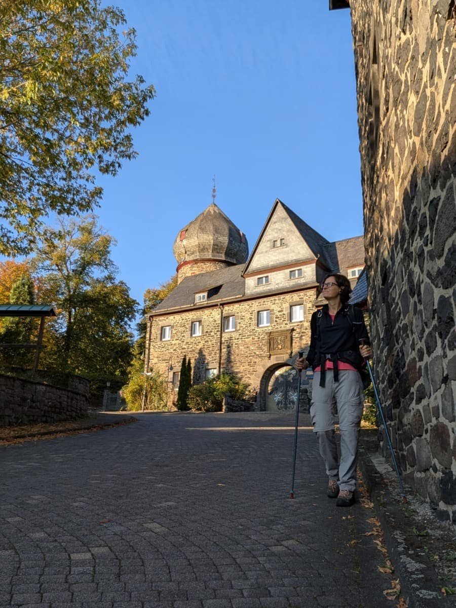 Linda steht mit Rucksack und Wanderstöcken auf der Straße vor Schloss Friedewald vor einem steinernen Gebäude mit rundem Turm.