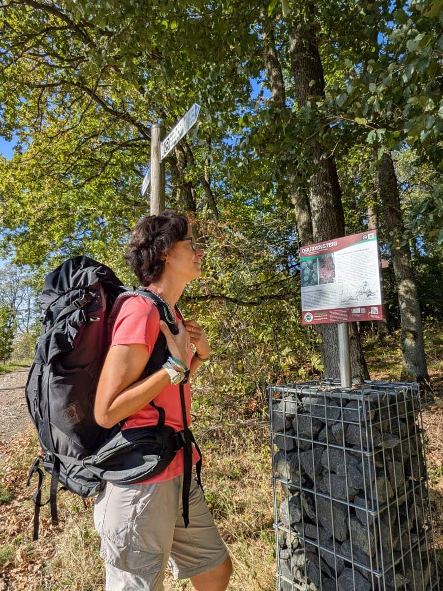 Linda steht mit großem Wanderrucksack neben einer Infotafel zum Druidensteig und einem Wegweiser am sonnigen Waldrand.