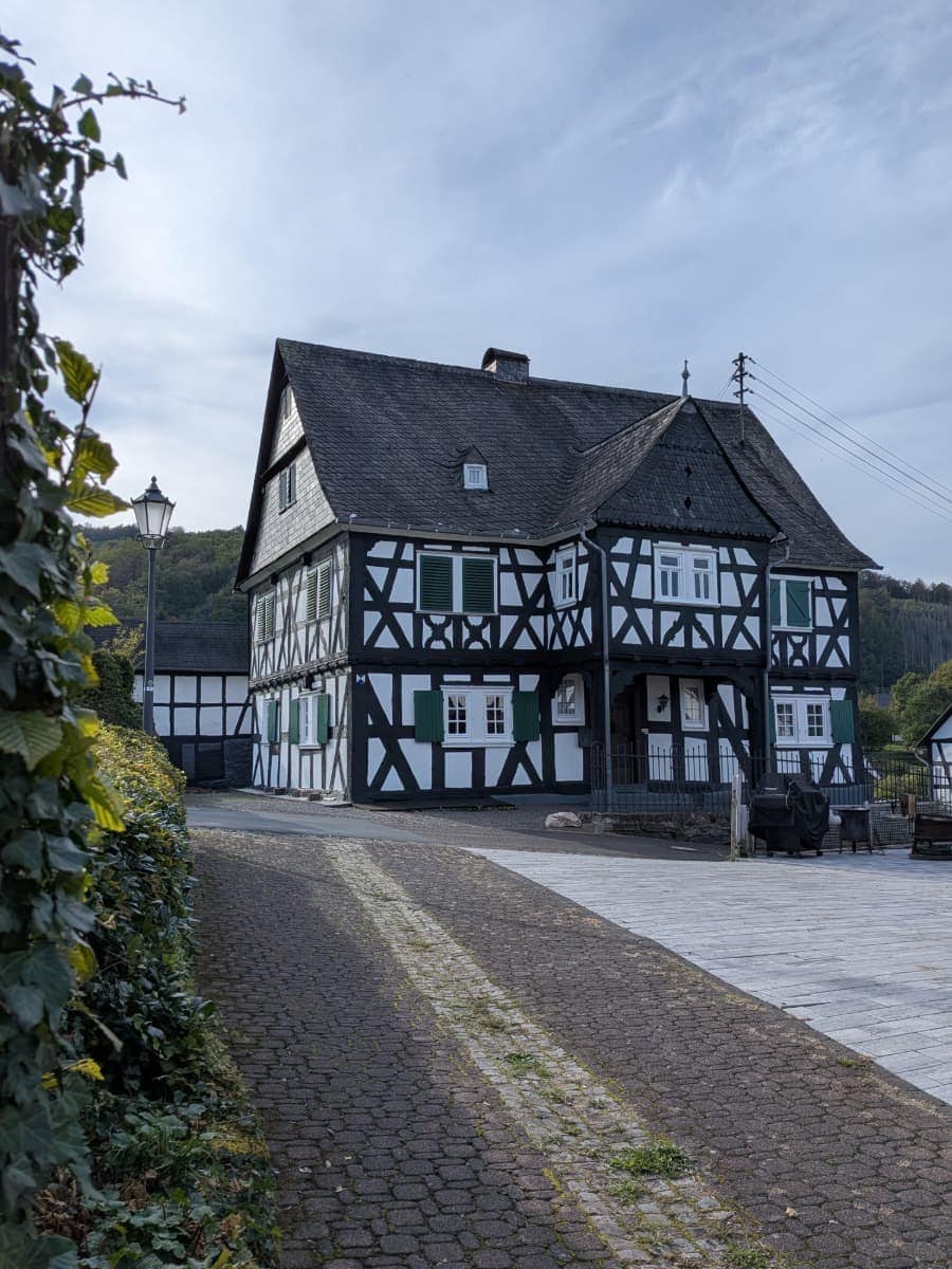 Das Hüttenschulzenhaus in Alsdorf hat schwarz-weißes Fachwerk, grüne Fensterläden und einen überdachten Vorbau an einer gepflasterten Straße.
